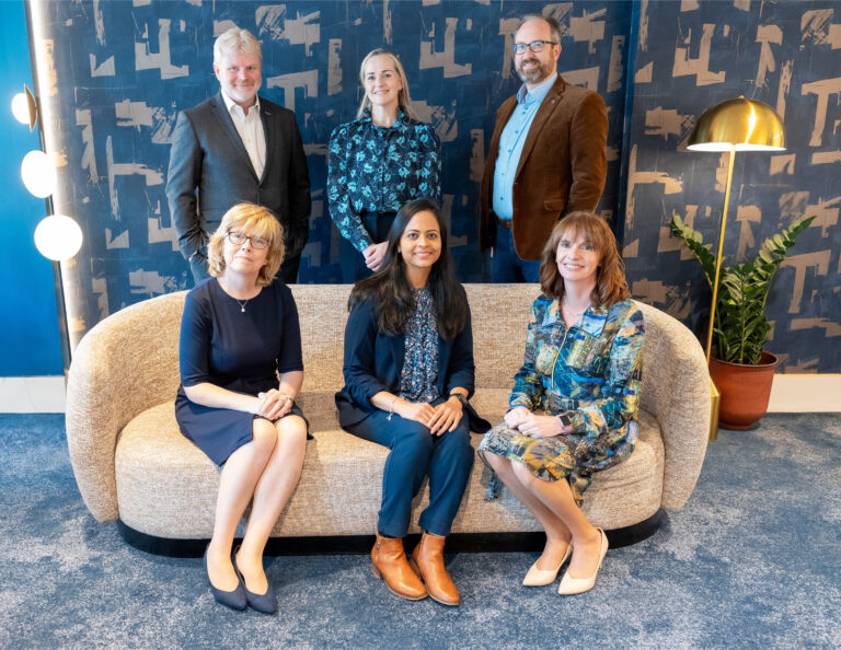 Six people posing for a group photo indoors; three standing behind a sofa and three seated on it. The setting features patterned blue wallpaper, a standard lamp, and a potted plant. All are dressed in business or smart-casual attire.