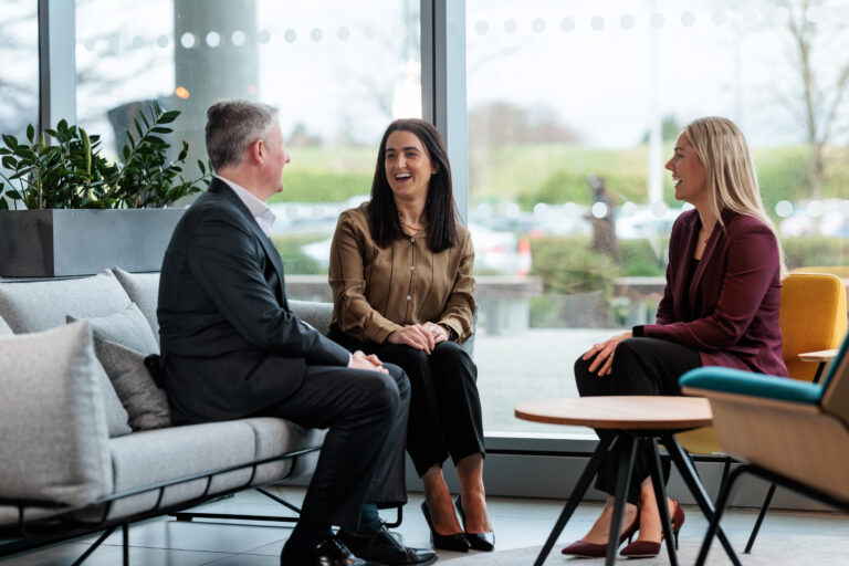 Three professionally dressed people sit on modern furniture by large windows, engaged in a lively conversation and smiling, with greenery and parked cars visible outside.
