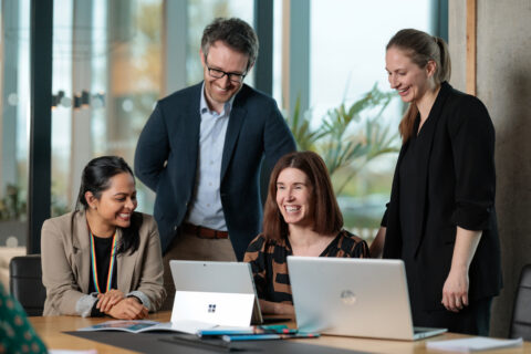 Four colleagues sit and stand around a table in a modern office, smiling and looking at two open laptops. Bright daylight streams through large windows, creating a collaborative and cheerful atmosphere.