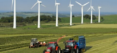 Three red tractors work in a green field harvesting crops, whilst several large white wind turbines stand in the background against a blue sky and rolling hills.