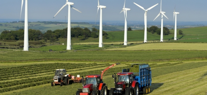 Three red tractors work in a green field harvesting crops, whilst several large white wind turbines stand in the background against a blue sky and rolling hills.