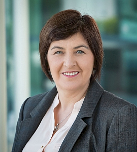 A woman with short brown hair wearing a grey blazer and white blouse smiles at the camera, standing in a bright indoor setting with a blurred background.