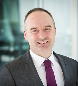 A man in a grey suit and purple tie is smiling at the camera. He has short brown hair and is standing in front of a blurred indoor background.