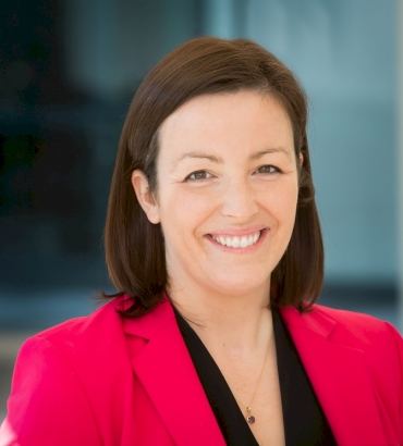 A woman with straight, dark brown hair smiles at the camera. She is wearing a bright pink blazer over a black top, standing in front of a softly blurred blue background.
