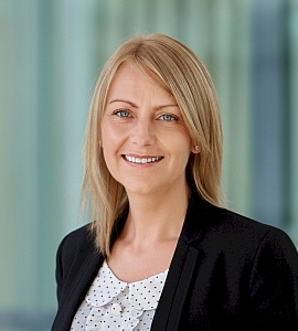 A woman with straight blonde hair wearing a black blazer and a white blouse with black polka dots, smiling in front of a blurred, light-coloured background.