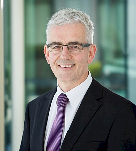 A man with short grey hair and glasses is smiling, wearing a black suit, white shirt, and purple patterned tie, standing in a modern office setting with glass windows in the background.