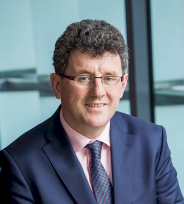 A man with curly dark hair, glasses, and a navy blue suit, light pink shirt, and patterned tie smiles at the camera in a modern office setting with large windows.
