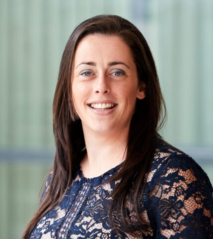 A woman with long brown hair, wearing a navy blue lace top, smiles at the camera against a blurred green background.