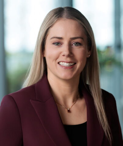 A woman with long blonde hair, wearing a maroon blazer and a necklace, smiles at the camera. The background is softly blurred with natural light and greenery visible.