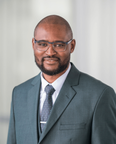 A man wearing glasses, a grey suit, white shirt, and a patterned tie is smiling softly while posing for a professional headshot against a light, blurred background.