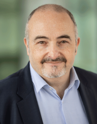 A middle-aged man with a bald head, grey beard, and moustache is wearing a dark blazer over a light blue shirt. He is smiling slightly and looking at the camera against a blurred green background.