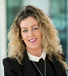 A woman with long, curly blonde hair is smiling and wearing a black blazer over a white collared shirt. The background is softly blurred, suggesting an indoor professional setting.