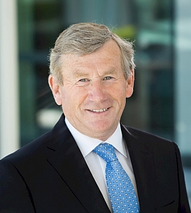 A middle-aged man with fair hair wearing a dark suit, white shirt, and blue polka dot tie smiles in front of a blurred office background.