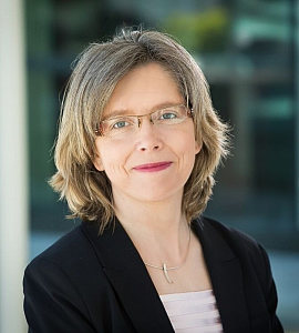 A woman with shoulder-length blonde hair and glasses, wearing a black blazer and a light-coloured top, smiles softly at the camera. The background is blurred with hints of greenery and building structures.