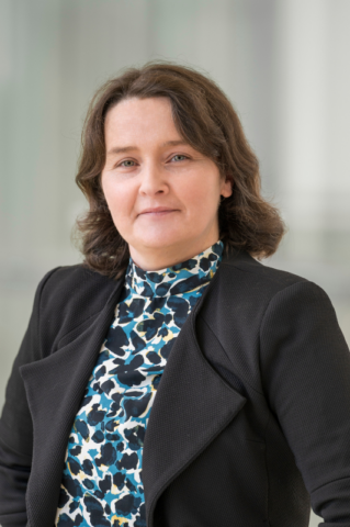 A woman with wavy brown hair, wearing a black blazer and a patterned high-neck blouse, poses for a professional portrait against a blurred light background.