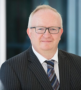 A middle-aged man with short, fair hair and glasses is wearing a dark pinstripe suit, white shirt, and a striped tie, smiling slightly while posing indoors with a blurred background.