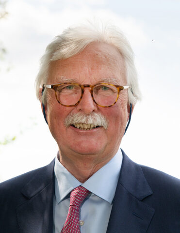 An older man with white hair and a moustache, wearing glasses, a dark suit, a light blue shirt, and a pink patterned tie, smiling outdoors with a bright sky in the background.