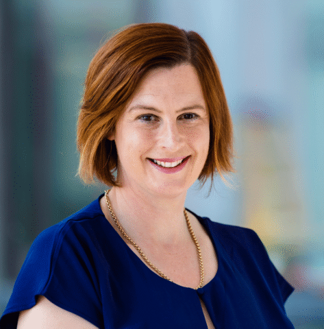 A woman with short, reddish-brown hair smiles at the camera. She is wearing a navy blue blouse and a gold chain necklace. The background is softly blurred in blue and grey tones.