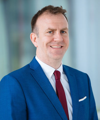A man in a blue suit with a white shirt and red tie stands smiling in front of a blurred indoor background.