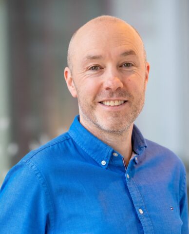 A smiling man with a shaven head and light facial hair, wearing a bright blue button-up shirt, stands indoors with a blurred background.