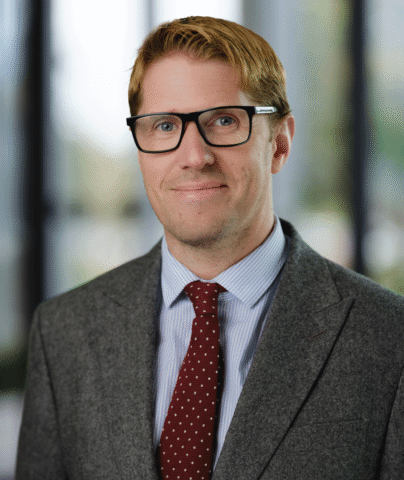 A man with light brown hair, wearing black glasses, a grey suit, a blue striped shirt, and a red tie with white spots, stands in front of a blurred office background, smiling slightly at the camera.