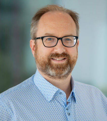 A man with light skin, glasses, and a reddish-brown beard is smiling. He is wearing a light blue patterned shirt and is posed in front of a blurred, neutral background.