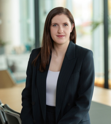 A woman with straight brown hair, wearing a dark blazer over a white top, is sitting in a modern office with glass walls and natural light. She is looking at the camera with a neutral expression.