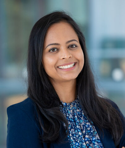 A woman with long dark hair smiles at the camera. She is wearing a navy blazer over a blue floral blouse, and stands in front of a blurred indoor background.