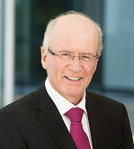 A smiling older man with glasses, short thinning grey hair, and wearing a dark suit, white shirt, and red polka-dot tie, stands in front of a blurred modern office background.