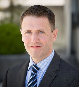 A man in a dark suit, light blue shirt, and striped tie is smiling softly outdoors. The background is blurred with greenery and a building visible.