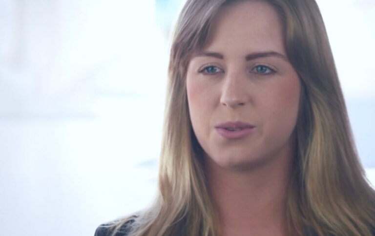 A woman with long, light brown hair and blue eyes is looking slightly to the side, speaking. She is indoors, with a soft, blurred white background.