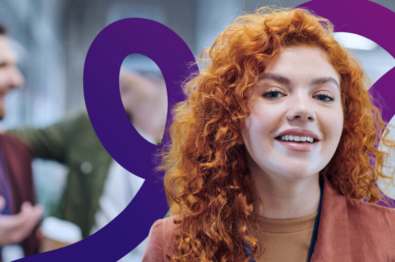 A young woman with curly red hair smiles at the camera. She wears a brown jacket and stands indoors, with two people blurred in the background and a purple decorative swirl in the foreground.