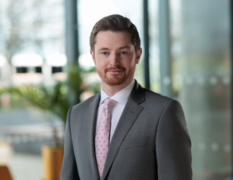 A man with short brown hair and a trimmed beard, wearing a grey suit, white shirt, and pink polka-dot tie, stands indoors in front of large windows with plants and blurred outdoor scenery in the background.