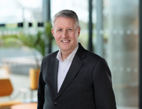 A smiling man with short grey hair wearing a dark suit and white shirt stands indoors in a modern, bright office setting with blurred background elements.