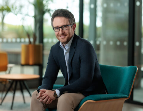 A man with glasses, beard, and dark hair sits on a modern teal chair, smiling at the camera. He is wearing a navy blazer, light shirt, and khaki trousers. The background shows large windows and indoor plants.