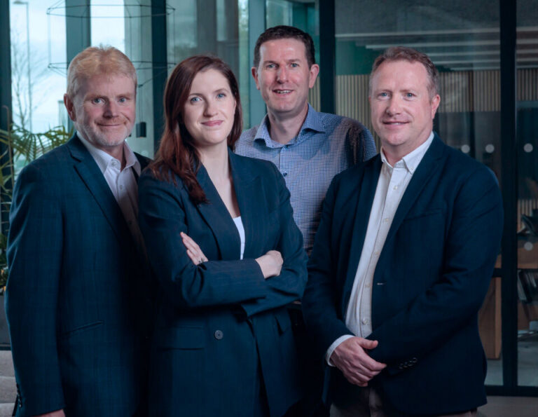 Four people wearing business attire stand together indoors, smiling at the camera. A woman with arms folded stands in front, with three men standing beside and behind her. The background shows a modern office setting.