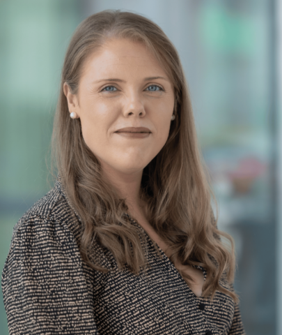 A woman with long light brown hair, blue eyes, and fair skin is wearing a patterned blouse and pearl earrings, standing in front of a blurred background with soft green and blue tones.