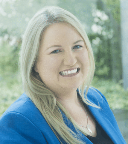 A woman with long blonde hair, wearing a blue blazer and a necklace, smiles at the camera with greenery and bright natural light in the background.