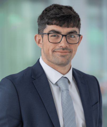 A man with short dark hair, glasses, and a trimmed beard is wearing a navy suit, white shirt, and light grey tie. He is smiling softly and standing in front of a blurred indoor background.