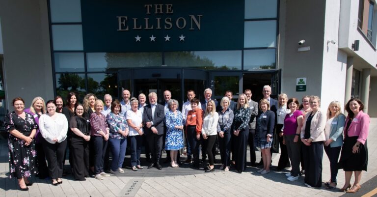 A large group of people pose for a photo outside the entrance of The Ellison hotel on a sunny day, standing in two rows and smiling at the camera.