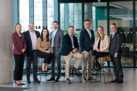 Eight professionally dressed adults pose and smile in a modern office space with large windows and contemporary furniture. Four are seated on tall stools, whilst the others stand beside them.