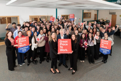 A large group of people smiling and holding red signs that read Great Place to Work. Certified 2024 in an office space, celebrating their workplace recognition.