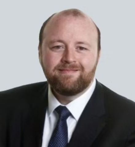 A man with a light complexion, brown hair, and a beard is wearing a dark suit, white shirt, and blue tie. He is smiling slightly and posed against a plain, light grey background.