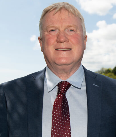 A middle-aged man with short fair hair, wearing a blue suit, light blue shirt, and red patterned tie, stands outdoors smiling with a blue sky and clouds in the background.