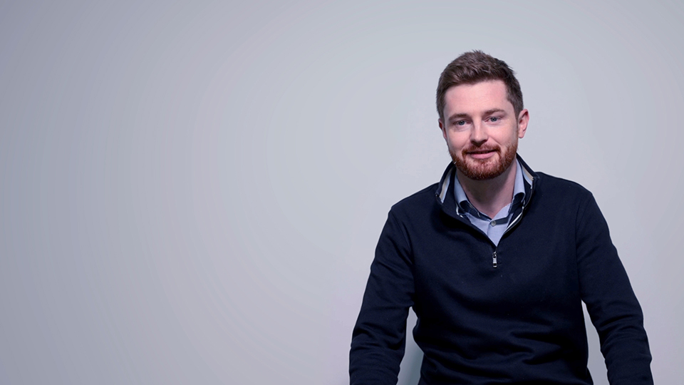 A man with short brown hair and a beard, wearing a navy jumper over a collared shirt, sits against a plain light grey background, smiling slightly.