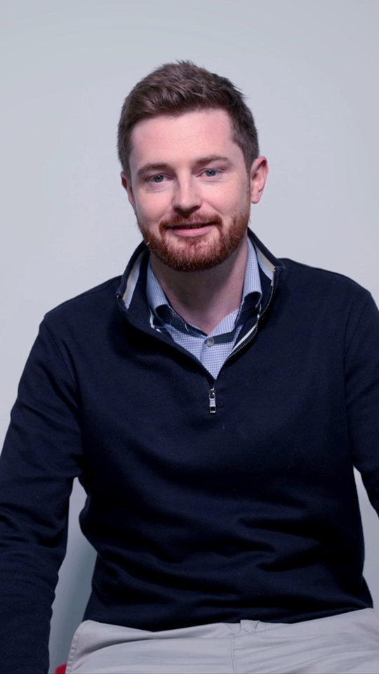 A man with short brown hair and a beard sits against a plain light grey background, wearing a navy quarter-zip jumper over a blue collared shirt and light trousers. He is smiling slightly and looking at the camera.