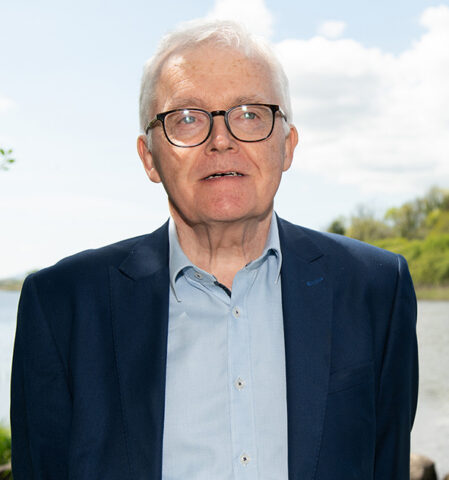 An older man with short white hair and glasses, wearing a blue suit jacket and light blue shirt, stands outdoors near a body of water with trees and a cloudy sky in the background.