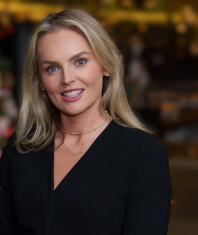 A woman with long blonde hair and blue eyes is smiling. She is wearing a black top and delicate gold necklaces, with a blurred indoor background behind her.