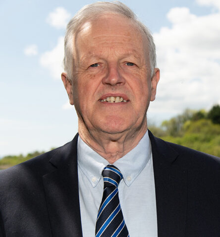 An older man with short grey hair wears a dark suit, blue shirt, and striped tie, standing outdoors with a partly cloudy sky and greenery in the background.