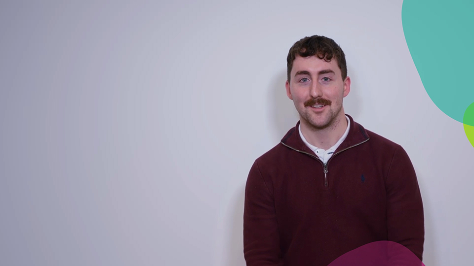 A man with short brown hair and a moustache, wearing a maroon jumper over a white shirt, sits against a light grey background with colourful abstract shapes on the right side of the image.